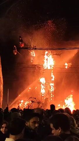 At night, a large crowd watches a tall, open-framed structure ablaze with fire. The structure is likely made of wood, with flames licking up its height. Two streams of water, possibly from fire hoses, are being directed at the fire. Power lines run above the scene. The crowd is made up of people of varying ages and genders, silhouetted against the bright flames. The overall environment is smoky, with a celebratory or ceremonial atmosphere.