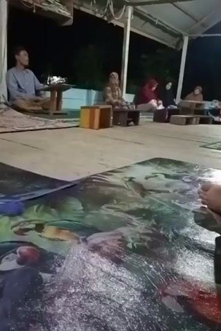 A gathering of people is taking place on a raised wooden platform outdoors at night. A man sits at one end of the platform, while a group of women, most wearing headscarves, are seated in a semicircle. The women are arranged around small, wooden stools or tables. A large, decorative mat lies in the foreground, partially obscuring the view of the group. The camera is positioned low to the ground, and the person holding the camera has their foot visible in the frame. The area is dimly lit by a single bulb, giving a slightly dark atmosphere to the scene.