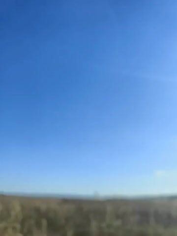 The video shows a wide shot of a rural landscape under a clear blue sky. The foreground is a field of harvested crops, with rows of stubble visible. In the middle distance, there is a small town with buildings and a tall, slender structure that resembles a water tower or silo. The horizon is flat, suggesting a vast plain or agricultural area. The overall impression is one of open space and agricultural activity. There are no people or animals visible in the video. The lighting suggests it is daytime, likely late morning or early afternoon, given the bright sky and the angle of the sun. The camera appears to be moving slowly, possibly from a vehicle, as indicated by the slight motion blur.