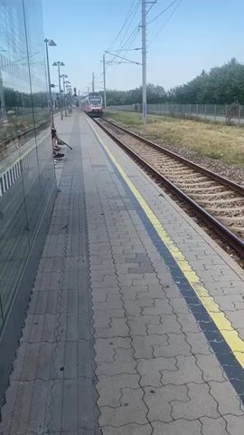 The video shows a train station platform with a train approaching on the tracks. On the left side of the frame, there is a building with a reflective glass facade and some people sitting on a bench. The platform itself is paved with gray interlocking bricks and has yellow and black safety lines. The train, which is white and pink, is moving towards the camera on the right track. In the background, there is a grassy area with trees and a clear blue sky. The scene appears to be during the daytime.