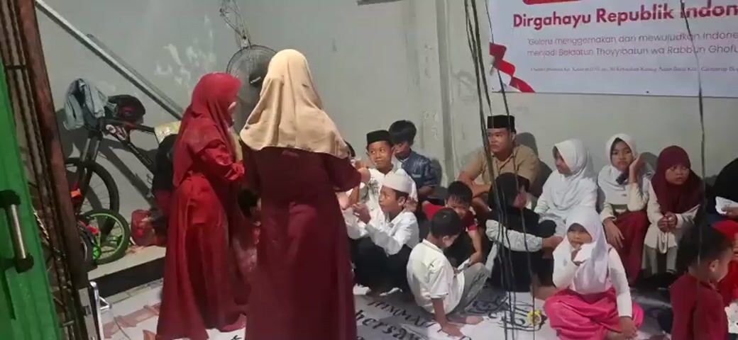 A group of people, mostly children and women, are gathered in a room. Two women in long red dresses and headscarves stand at the left, facing the others. A banner with Indonesian text and the Indonesian flag is visible in the background. The people are sitting on the floor, some are listening attentively, while others seem to be interacting. There is a fan on the left side of the room. The lighting is dim.