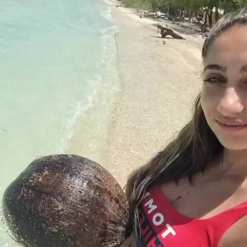 A young woman stands on a white sandy beach with clear turquoise water behind her. She is holding a brown, fibrous coconut in her left hand. She is wearing a red and blue swimsuit and has long brown hair. She smiles at the camera. In the background, a distant coastline can be seen with a mountainous range. The sky is partly cloudy.