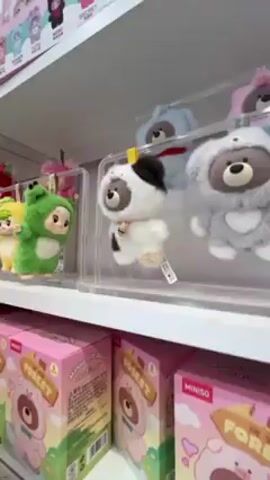 The camera pans across a shelf of small plush figures in clear plastic cases. The first few are bear-like characters, and then they transition to various cartoon characters such as My Melody, Hello Kitty, and a panda. The shelves below contain pink boxes with cartoon designs.