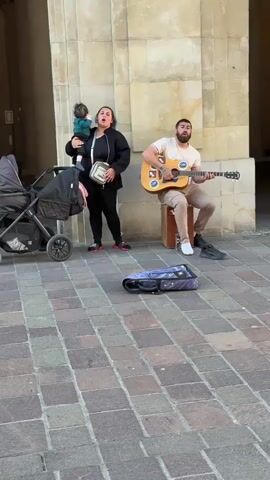 A street musician is busking under a stone archway in what appears to be a public square or passage. A man with a beard and tattoos, wearing a beige t-shirt and light brown pants, is seated on a wooden box, playing an acoustic guitar and singing. His left shoe is off, revealing a white sock, while his right foot wears a dark sneaker. His guitar has several stickers, including a peace sign.

To his left stands a woman with dark hair, dressed in a black long-sleeved top, black leggings, and pink and black sneakers. She is holding a baby wearing a green long-sleeved top and dark pants. The woman has a white crossbody bag with a "PUMA" logo. A dark-colored baby stroller is parked nearby on the far left.

An open dark blue or purple guitar case lies on the paved ground in front of the man, presumably for tips. The background features light-colored stone walls and a large archway, with an overhead light fixture visible further inside. A faint silhouette of another person can be seen in the distant archway. The ground is paved with reddish-brown and gray rectangular bricks. The scene is well-lit, indicating daytime. Both the man and woman appear to be singing or vocalizing, and the baby is looking around.