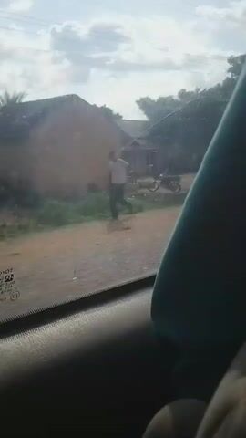The video shows a view from inside a car, looking out the passenger window. The exterior appears to be a dirt road with buildings and trees. In the foreground of the window, part of a car's interior is visible, including a black door panel and a turquoise headrest. The outside scenery passes by. There are several people visible in the distance, some standing near vehicles and structures that appear to be shops or stalls. One shot shows a white van with its trunk open. Another shot shows a person in a blue garment standing next to a vehicle. The sky is cloudy. The camera appears to be moving, suggesting the car is in motion.