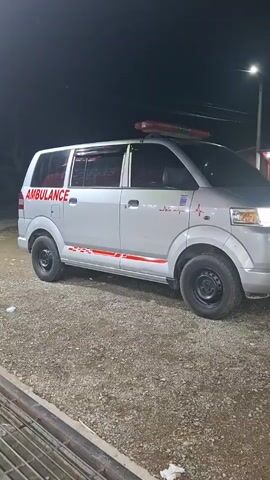 The video features a silver Suzuki APV van, modified as an ambulance, parked outdoors at night on a gravel surface. The vehicle displays "AMBULANCE" in red lettering on its side and front, along with a red heartbeat graphic on the front fender. Red decorative stripes are visible along the lower side of the vehicle. A red light bar is mounted on the roof, and blue and yellow flashing lights are active on the front bumper. The vehicle's headlights are illuminated. In the background, a structure with white framing and a red roof is partially visible. An engine sound is audible.
