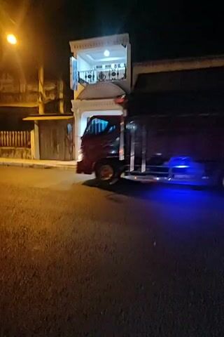 A truck with blue undercarriage lights drives down a street at night. A street light shines brightly overhead. A building with illuminated windows is visible to the right. Another truck is partially visible in the foreground, also at night.