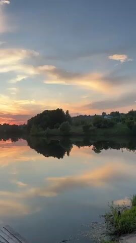 The video captures a serene sunset over a calm body of water. The sky is painted with soft hues of orange, pink, and blue, with wispy clouds reflecting on the water's surface. The reflection creates a mirror image of the sky and the silhouetted treeline on the opposite bank. A small wooden dock or pier is visible in the foreground on the left, with a section of wooden planks. On the right side of the dock, a small, green-roofed gazebo stands, equipped with benches and a table. Two life preservers are mounted on the railings of the gazebo. A small blue boat is moored to the left of the dock. The environment appears to be a tranquil natural setting, likely a lake or river, with a gentle incline of trees and houses on the distant shoreline.