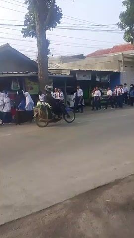 The video shows a group of students in uniform walking along a street. Some are walking in a line, while others are scattered. There are buildings and trees on the side of the street. A few motorcycles are also visible, with some people riding on them. The weather appears to be clear.