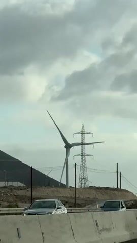 A vertical, eye-level shot shows a road with a white concrete barrier in the foreground. A silver car drives past on the road. Beyond the road, a landscape of dry, brown hills stretches out under a cloudy sky. A large wind turbine with three blades stands prominently on one of the hills, with a tall, lattice electrical pylon behind it. Other wind turbines are visible in the distance on other hills. A white van drives past on the road. A person is visible standing near the base of the large wind turbine. The scene appears to be an open, industrial landscape focused on renewable energy.