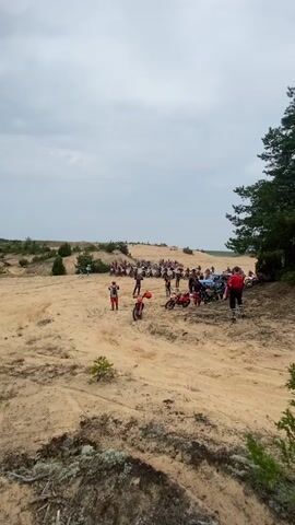 A group of motorcyclists ride their dirt bikes through a sandy, hilly terrain. There are numerous trees and bushes scattered throughout the landscape. The sky is cloudy. Many of the motorcycles are red, while others are white, yellow, and blue. Several people are standing around near the motorcycles, observing the riders. The riders are wearing helmets and protective gear. The ground is covered in sand and some patches of dry vegetation. Some of the motorcycles are kicking up dust as they ride.