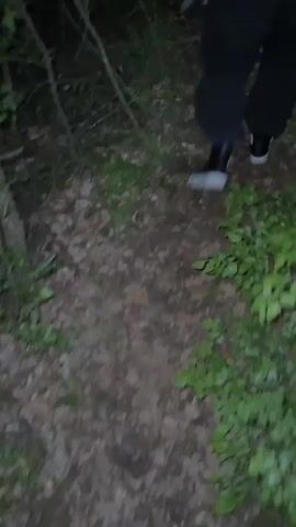 A person wearing dark pants, black and white shoes, and a purple plaid shirt with a black backpack is walking on a dirt path in a forest at night. The camera follows them from behind as they walk. The forest is dark, with sparse foliage and many small trees. The ground is covered in dead leaves.
