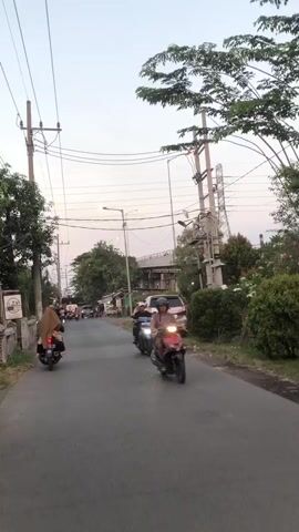 The video shows a street scene. Two people are riding motorbikes on a paved road. The person on the left is wearing a headscarf and riding a dark-colored motorbike. The person on the right is wearing a light-colored headscarf and riding a blue motorbike. A silver van is parked on the side of the road. There are trees and power lines along the street. The sky is visible, and it appears to be daytime, possibly late afternoon or early evening due to the lighting. The environment appears to be a rural or suburban area.