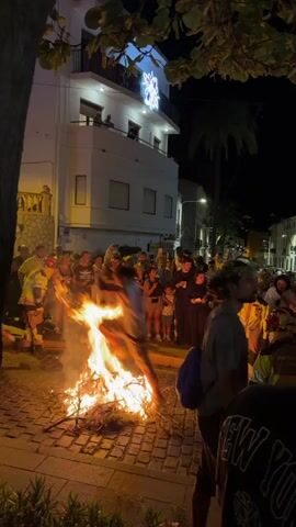 The video shows a nighttime street scene where a large bonfire is burning in the center of a cobblestone street. A crowd of people, appearing to be onlookers, are gathered around the bonfire, many holding up their phones to record. Some individuals are closer to the fire, with one person, dressed in light-colored clothing, captured mid-leap over the flames. Another person, wearing a white dress, is seen near the fire on the right side of the frame. Buildings with lit windows line the street, and decorative lights, including a snowflake motif on a balcony, are visible on one of the buildings. Palm trees and foliage are present in the foreground and background, suggesting a warm climate. The atmosphere appears to be celebratory, likely a festival or event.