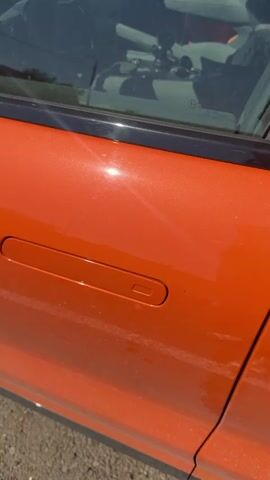 A close up shot of an orange car door. A person's finger with painted nails points at the door handle. The door then opens, revealing the interior of the car. The car appears to be parked outdoors on a dirt road with grass and some distant buildings visible.
