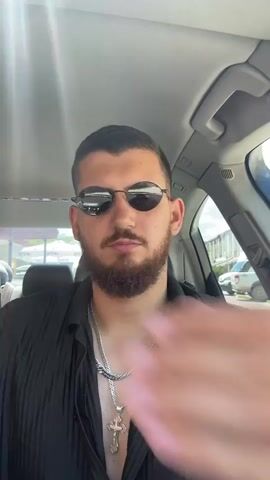 A man with short dark hair and a beard wearing sunglasses and a black collared shirt is sitting in the driver's seat of a car. He has on a silver watch, several silver necklaces, one with a cross pendant, and a silver ring. He is holding his hand over his mouth and then moves it away, revealing his face as he smiles. The interior of the car is visible, as is a bright, sunny outdoor scene through the windshield.