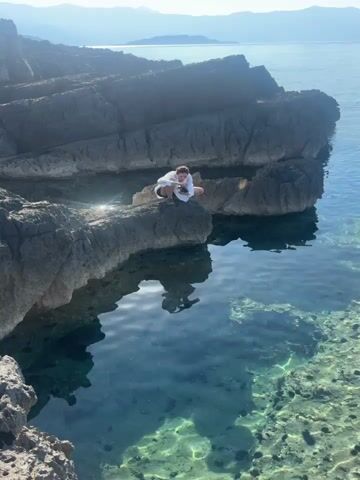A person crouches on a rocky outcrop overlooking clear blue water. The person is wearing a white shirt and dark shorts. The rocks are layered and rough. The water is shallow and appears to have a sandy bottom. In the distance, there are mountains or hills. The sky is bright and sunny.