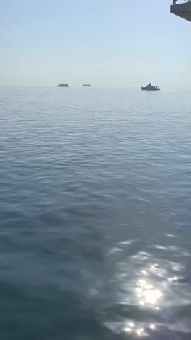 The video shows a wide expanse of calm, blue water under a clear, pale sky. In the distance, three boats are visible on the horizon. The closest boat on the right appears to be a smaller vessel, while the two boats further to the left are larger and have a more imposing presence. Sunlight glints off the surface of the water, creating bright reflections. The perspective is from a slightly elevated viewpoint, suggesting the camera is on a dock or a larger vessel. The overall impression is one of a serene and open seascape.