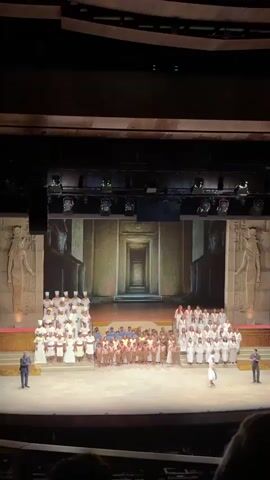 A stage presentation is underway, showcasing a large ensemble of performers dressed in historical costumes, likely representing ancient Egyptians.  The backdrop consists of a projected image of a temple corridor, flanked by large statues. A man in a suit stands to the left side of the stage, possibly an announcer or director, while another man in a suit stands to the right. The performers are arranged on tiered platforms. The audience is partially visible in the foreground. The venue appears to be a theater or auditorium.