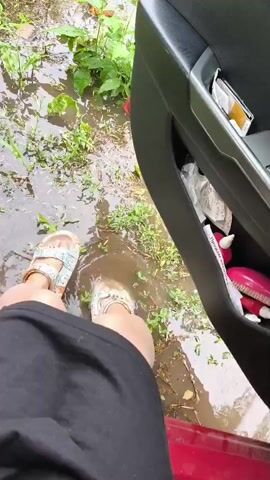 A person wearing sandals and a black skirt stands on a wet, muddy ground next to an open car door. The ground is covered in puddles and green vegetation. The car door pocket contains various items including a yellow packet, some white wrappers, and a pink object with white accents. The person appears to be stepping out of the car and into the water.