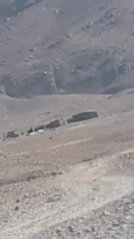 The video depicts a mountainous, arid landscape. Initially, a blurry view of some structures is visible on the hillside. The camera angle shifts, revealing a view of the mountain from a road. The mountain is brown and barren, with a few small trees scattered on the slope. The sky is clear and blue.