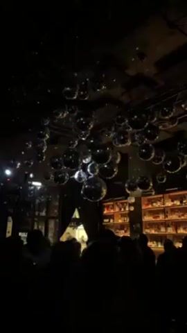 The video shows the interior of a dimly lit bar or club.  The foreground is filled with silhouettes of people, suggesting a crowded space. Above the crowd, numerous disco balls hang from the ceiling, reflecting light and creating a sparkling effect. In the background, a well-stocked bar is visible with shelves filled with bottles. There appear to be more people standing near the bar. The overall atmosphere is festive and energetic.