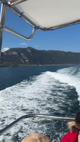 The video begins with a view from a boat moving on the water, leaving a frothy white wake. The camera is angled downwards and towards the right, showing the wake and the side of the boat. A person in a red shirt is visible in the lower right corner, facing away from the camera. The boat appears to have a white canopy and a metal railing. In the distance, a shoreline is visible with green, tree-covered mountains and a cluster of buildings. The sky is clear and blue. The camera pans slightly to the right, revealing more of the distant shoreline and mountains. The boat continues to move forward, creating a significant amount of white foam in its wake. The mountains in the background are large and imposing, with some areas appearing rocky. The water is a deep blue. The video continues to show the boat moving through the water, with the wake as a prominent feature. The shoreline remains visible in the distance, and the sky is consistently blue. There are no other people clearly visible in the frame besides the partial view of the person in the red shirt. The overall impression is of a sunny day spent boating on a large body of water with mountainous scenery.