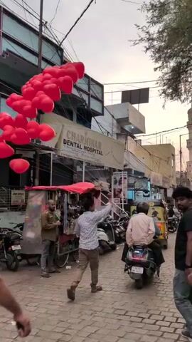 The video displays a street-level view of a bustling urban thoroughfare. A large cluster of approximately 30-40 red heart-shaped balloons floats upwards and drifts towards the background. A man in a light-colored checkered shirt runs towards the background, looking up and gesturing towards the balloons. On the left, a building has a prominent sign for "CHARMINAR DENTAL HOSPITAL" and another reading "CHARMINAR." Below the signs, a street vendor operates a cart with a red canopy. Multiple pedestrians walk in various directions, some towards the camera and others away. Several motorbikes are parked along the street or are in motion. In the distant background, a large, ornate stone archway with a tall minaret-like structure is visible against a dusky sky. Overhead power lines crisscross the frame, and a street light is illuminated.