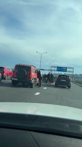 A group of motorcyclists appear to have stopped on the side of a highway. A red SUV is parked behind them, and another car is visible further down the road. A red triangular warning sign is placed on the road in front of the motorcycles. One person, wearing a black vest over a jacket and pants, is standing next to the red SUV and appears to be interacting with someone inside the vehicle. Other individuals in motorcycle gear are standing with their motorcycles. The sky is cloudy, and overhead signs indicate an exit or highway direction.