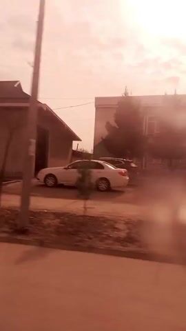 The video, shot from a moving vehicle, shows a roadside view under a bright, hazy sky. The camera focuses on various buildings as they pass by. These buildings are primarily single-story structures with a mix of flat and pitched roofs, predominantly painted in light, neutral tones. One building features a yellow entrance and another displays a sign reading "OQTEPA" or something similar in Cyrillic script. Patches of dry, unpaved ground and sparse vegetation border the road. The overall color cast of the video is warm, likely due to the time of day or atmospheric conditions. There is some slight distortion visible, potentially caused by water on the windshield of the vehicle.