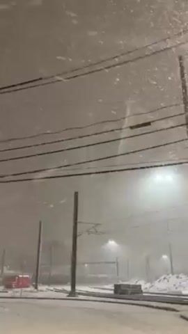 The video shows a snowy scene at night. Large snowflakes are falling heavily, creating a dense whiteout effect. In the foreground, there are railway tracks covered in snow, with overhead power lines and supporting poles visible. Streetlights illuminate the area, creating a bright glow amidst the snowfall. A red structure is partially visible on the left. A car covered in snow is parked on the right side.