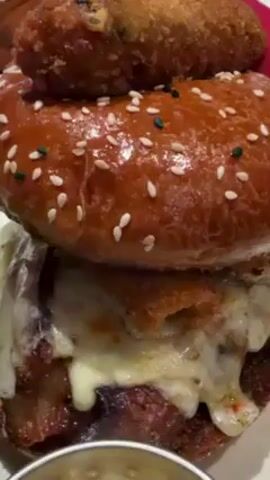 The video shows a close-up of a tall burger with a crispy topping on top, possibly at a restaurant. The burger has a glossy, sesame-seed covered bun, with melted cheese, bacon, and some other fillings in between. A small fried roll-shaped item is balanced on top of the bun.  A wooden pick with the words "Gordon Ramsay Burger" is sticking out of the top bun. The burger sits on a white square plate, with small metal containers of condiments next to it. Other burgers and glasses of water are visible in the background, suggesting the scene is at a restaurant. People are indistinctly visible in the background.