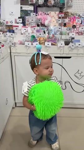 A toddler wearing a striped shirt and blue jeans stands in a store aisle, holding a large, neon green spiked ball toy. The toddler has a blue, tentacle-like hair accessory on their head. The background shows shelves stocked with various merchandise, including small bags, plush toys, and stationery. The store displays white cabinets with black line drawings on them, including a camera, a crown, and the words "LIKE SHARE". The toddler turns their head away from the camera and walks towards the right side of the frame, still clutching the toy.