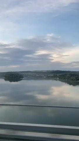 The video presents a serene, scenic view of a wide river from the perspective of a moving vehicle, likely a car or train. The river reflects the sky above, creating a mirror-like surface.  There are small, wooded islands or peninsulas visible in the river. The sky is partly cloudy, with a mix of blue sky and white and gray clouds, suggesting a late afternoon or early morning light.  The horizon line features a blend of green hills and distant structures, possibly a town or village.  A metal railing is visible at the bottom of the frame, indicating the video was taken from a bridge or elevated structure. The overall tone is tranquil and peaceful, focusing on the natural beauty of the landscape.
