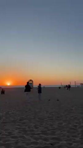 The video depicts a beach at sunset. The sun is setting over the ocean, casting a warm orange glow across the sky. The beach is sandy and populated with several people. Two figures stand near a yellow playground structure, possibly taking photos or observing the sunset. Other individuals are seen walking or sitting on the beach, and a volleyball net is visible in the distance. The overall scene is tranquil, capturing a moment of appreciation for the natural beauty of the setting sun.