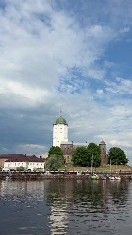 A body of water in the foreground reflects a castle situated on a small landmass in the background. The castle has a prominent white tower with a green dome and a flag at its peak, and a darker stone section with a smaller tower. Green trees surround the castle. To the left of the castle are white buildings with dark roofs. Sailboats are docked to the right of the castle. The sky above is a mix of bright blue with scattered white clouds and a darker, cloudier section on the left.