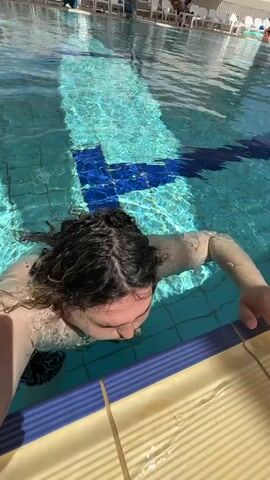 The video shows a fair-skinned man with dark hair and a beard in a swimming pool. The pool appears to be located outside, surrounded by lounge chairs and shade structures. The man addresses the camera, identifying himself and the pool. He winks at the end.