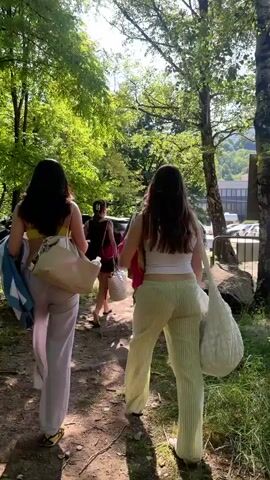The video shows three young women walking away from the camera through a park-like area. The first two women are in the foreground, wearing light-colored pants and carrying bags. One has a towel slung over her shoulder. The third woman is slightly behind and wearing a short dress. The setting includes trees, grass, and a fence, leading to a parking lot filled with cars. A building can be seen in the background against a backdrop of hills. The sun is shining, creating a bright, daytime scene.