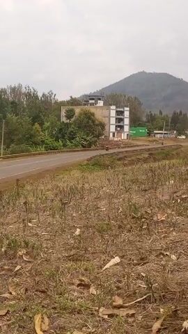 The video shows a view of a road running through a rural area. On one side of the road is a dry field, while on the other side is a building under construction surrounded by trees. In the distance, a hill or mountain can be seen under a cloudy sky. There is a person walking on the road in the direction of the viewer.