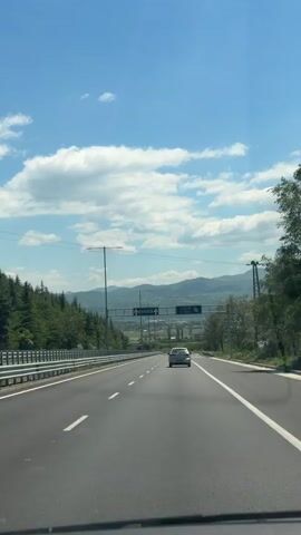 The view from the front of a car driving on a highway in Bulgaria. A silver car drives ahead in the distance. Green trees and hills line the sides of the highway, and mountains are visible in the background under a blue, partly cloudy sky. Road signs indicate directions to “Kulata” and “Riltsi b. pole”. The highway is well-paved with white dividing lines.