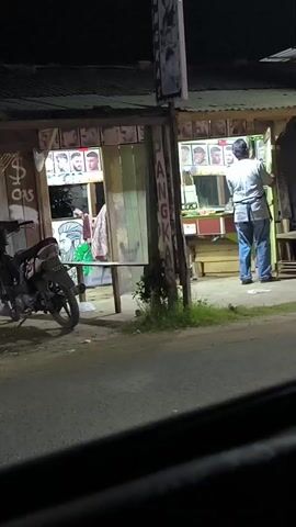 A static shot captures a barber shop at night. The shop appears to be made of wood with a corrugated metal roof. Inside, several framed pictures of hairstyles are displayed along the top of the shop. A motorcycle is parked to the left of the shop. To the right, a man wearing a light-colored shirt and blue jeans, with an apron tied around his waist, stands facing away from the camera, engaged in an activity inside the shop. The shop is illuminated by artificial light, casting shadows on the surrounding area. The street in front of the shop is visible, as is a portion of a vehicle from which the video is likely being recorded.
