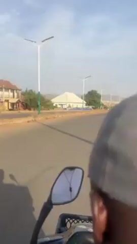 The video shows a person riding a motorcycle on a paved road. The rider is wearing a grey baseball cap and a white shirt. The road is lined with modern streetlights. In the background is a building with a large, white roof, and beyond that, a distant, hilly landscape. The sky is light blue. The perspective is from behind the rider, focusing primarily on the road ahead and occasionally on the rider's face and surroundings.