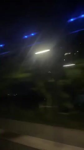 The video captures a night scene from inside a moving vehicle, likely a bus, given the blue lights visible on the ceiling. The perspective is through a window, looking out at the passing environment. Outside, the scene is dark, illuminated primarily by artificial lights. Streaks of light from streetlights or other vehicles are visible, indicating motion. In the distance, faint clusters of lights suggest buildings or a cityscape. Around the 0:00:03 mark, a large, illuminated billboard is briefly seen, but its text is illegible due to motion blur and distance. The overall impression is of a late-night journey. No people are visible. No legible text is present in the video.