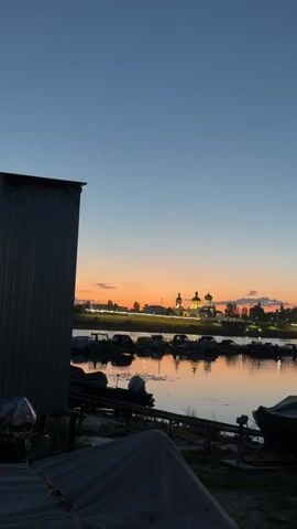 A video pans across a waterfront at dusk. In the foreground, several boats are pulled onto the shore, some covered with tarps. A large, grey, industrial-style building is visible on the left. The middle ground features a body of water with several smaller boats moored. Across the water, a town or city skyline is silhouetted against a colorful sunset sky. Prominent on the skyline are the domes of churches or cathedrals, illuminated. Trees and streetlights are also visible along the shoreline. The sky transitions from a deep blue overhead to shades of orange, pink, and yellow near the horizon. The water reflects the colors of the sky and the lights of the distant town. The video appears to be a static shot with a slight pan or zoom effect.