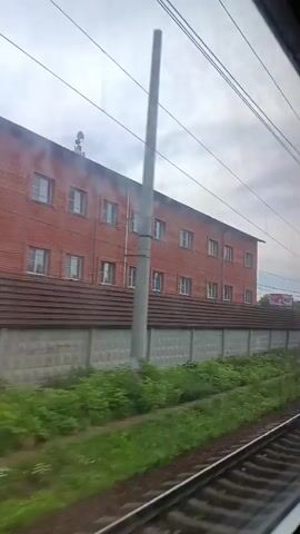 The video shows a view from a moving train. On the left is a red brick building with multiple windows. A grey concrete fence with a tiled top runs along the side of the building. Green grass and vegetation grow alongside the fence and the train tracks. Overhead, power lines and a utility pole are visible. In the distance, there appear to be more buildings, trees, and a billboard. The sky is overcast. The train tracks are in the foreground.