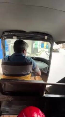 The view from the back seat of a three-wheeled vehicle, likely a tuk-tuk or auto rickshaw. A man with short graying hair wearing a yellow, blue, and gray striped shirt is driving. The tuk-tuk is traveling on a narrow street with other vehicles and buildings visible through the windshield. A red helmet sits on the back seat. The tuk-tuk is emitting a high-pitched squeal.
