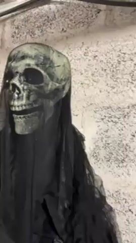 A close-up shot shows a static Halloween decoration resembling a grim reaper. The decoration consists of a gray skull with dark eye sockets and visible teeth, covered with dark, flowing fabric. The background is a textured off-white wall, slightly out of focus. The camera angle provides a direct view of the skull, with slight panning movements.