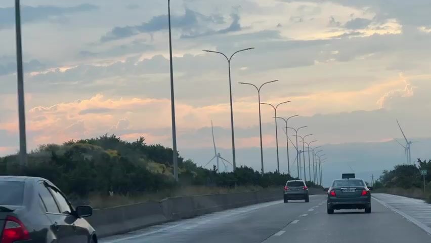 The video captures a view from a vehicle driving on a highway. Several cars are visible on the road, including one in the foreground on the left with a license plate that appears to read "DT 770 TT". The sky is cloudy with streaks of orange and pink, suggesting either sunrise or sunset. Tall streetlights line the right side of the highway. In the distance, wind turbines are visible against the horizon. The landscape on the left side of the highway is covered in dense green vegetation.