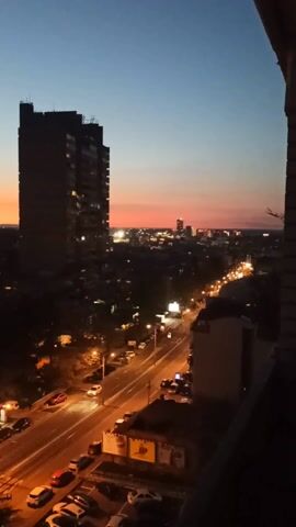 Here is a description of the video:

The video shows a cityscape at dusk. The sky transitions from a deep blue at the top to a blend of orange and pink near the horizon. In the foreground, there's a tall, dark building on the left. Streets lined with streetlights and cars are visible below. Some buildings and trees surround the streets. The video pans slightly to the left, revealing more of the cityscape and a park-like area. The overall scene has a quiet and urban feel.