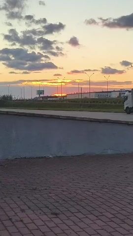 The video captures a sunset with vibrant orange and yellow hues streaking across the sky, interspersed with dark clouds. A road with streetlights runs horizontally across the middle ground, with a concrete barrier in the foreground. In the distance, there are industrial buildings and a billboard. A white truck is visible on the road, moving from right to left. The ground in the foreground is paved with reddish-brown bricks.